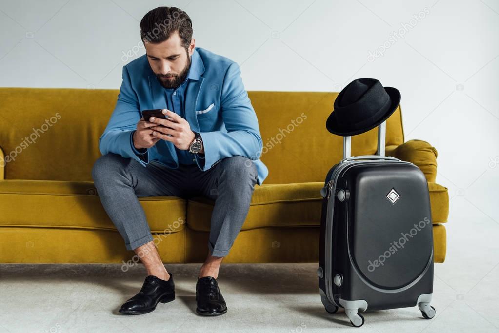 handsome stylish businessman using smartphone and sitting on sofa with luggage 