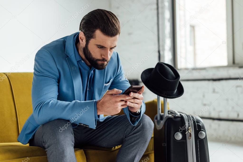 concentrated businessman using smartphone and sitting on sofa with travel bag