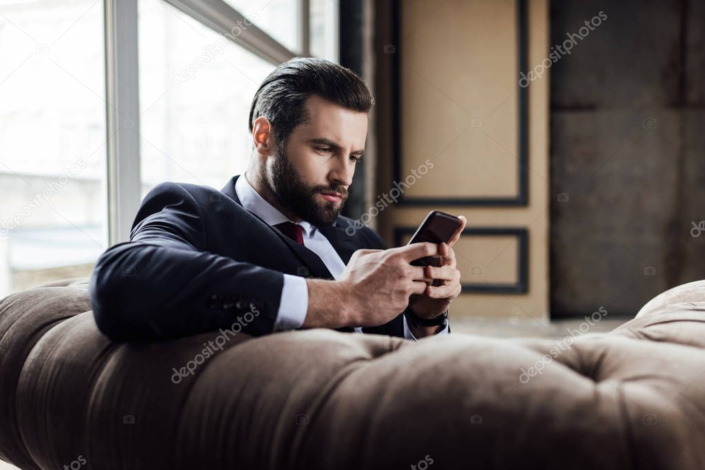 confident bearded businessman using smartphone and sitting in armchair 