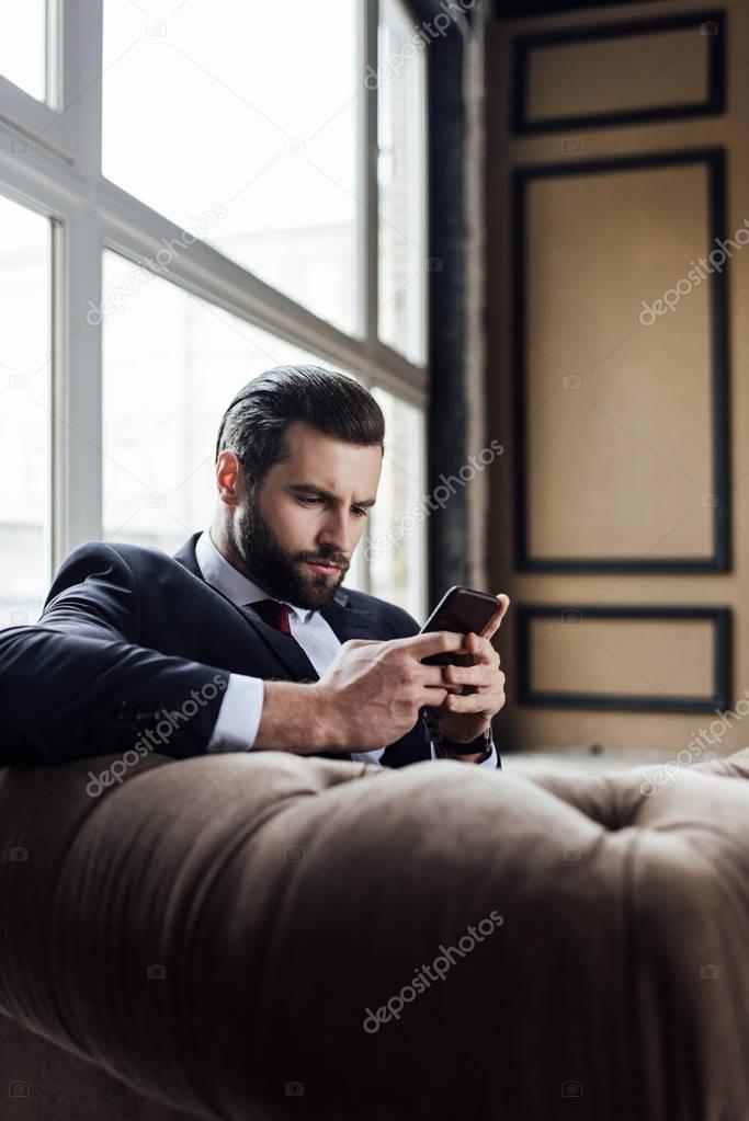 fashionable bearded businessman messaging on smartphone while sitting in armchair 
