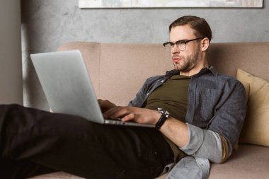 ciddi genç adam kanepede dizüstü bilgisayar ile çalışma