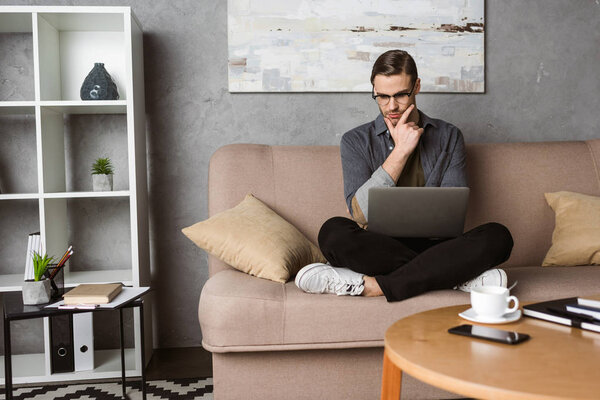 young thoughtful freelancer working with laptop while sitting on couch