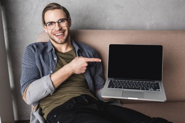 genç gülümseyen adam kanepede oturan ve dizüstü bilgisayar ekranında işaret