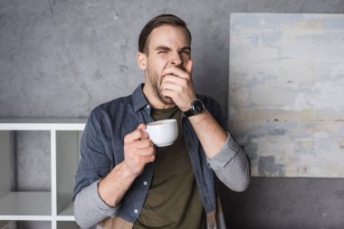 exhausted yawning young man with cup of coffee clipart