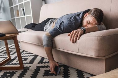 exhausted young man sleeping on couch with smartphone in hand clipart
