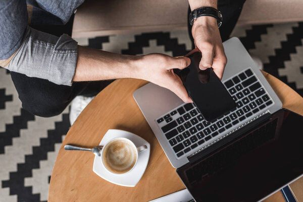 cropped shot fo man using smartphone with laptop on coffee table