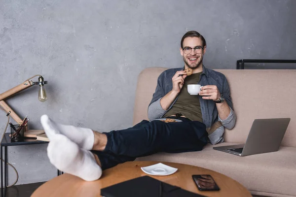 年轻英俊的人与笔记本电脑喝咖啡与饼干在沙发上 — 图库照片