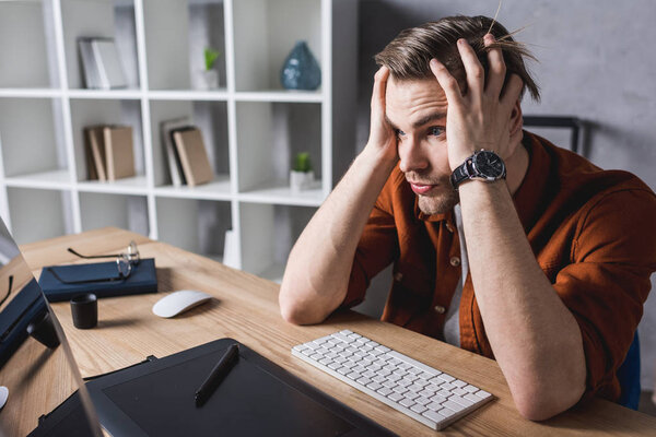 bewildered designer sitting at workplace and holding head with hands
