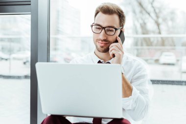 smartphone üzerinde konuşmak ve modern ofis penceresinde, dizüstü bilgisayar kullanarak yakışıklı işadamı