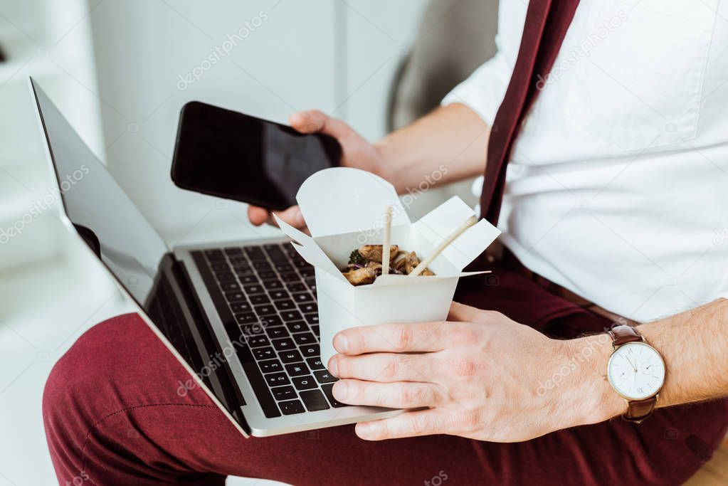 cropped view of businessman holding box with noodles, smartphone and laptop
