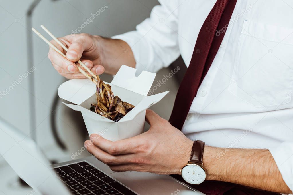 cropped view of businessman with laptop eating noodles in office