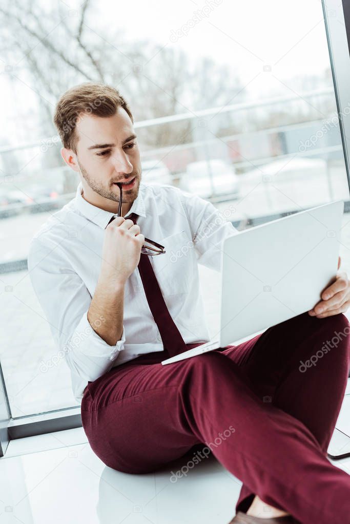 handsome pensive businessman using laptop and sitting on floor