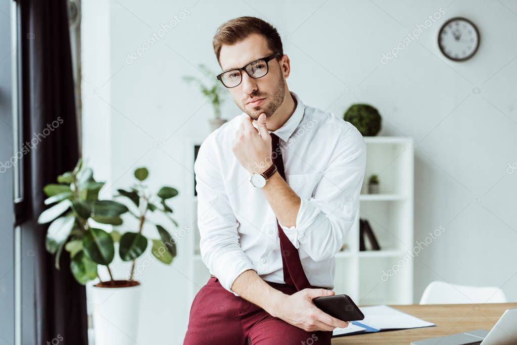 handsome pensive businessman in eyeglasses holding smartphone at workplace