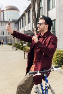 şık adam gösteren zafer işareti gülümseyen ve bisiklet üzerinde eğilerek smartphone selfie alarak