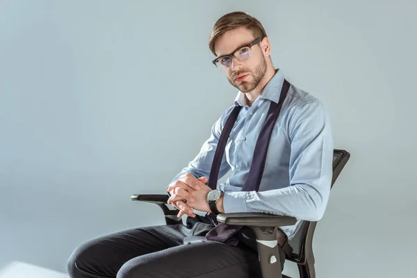 Joven empresario confiado sentado en silla con las manos apretadas, aislado en gris - foto de stock