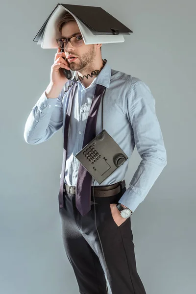 Confused businessman holding notebook on his head while talking on phone isolated on grey — Stock Photo