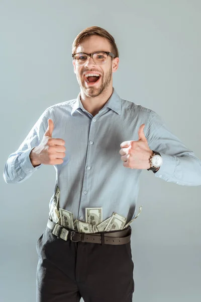 Молодой возбужденный бизнесмен с долларовыми купюрами и штанами показывает большие пальцы, изолированные на сером — стоковое фото