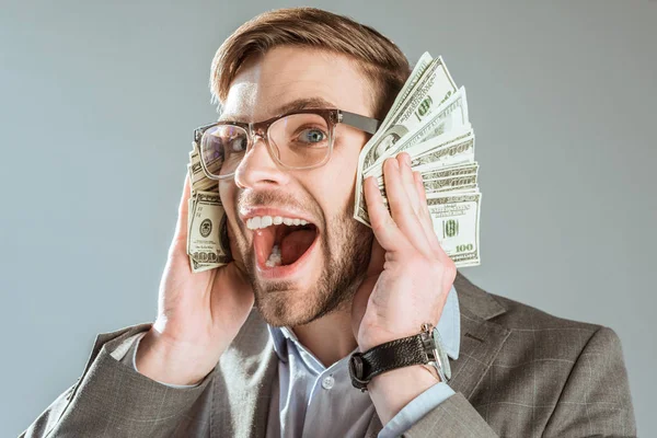 Rico homem de negócios feliz segurando notas de dólar perto de seu rosto isolado em cinza — Fotografia de Stock