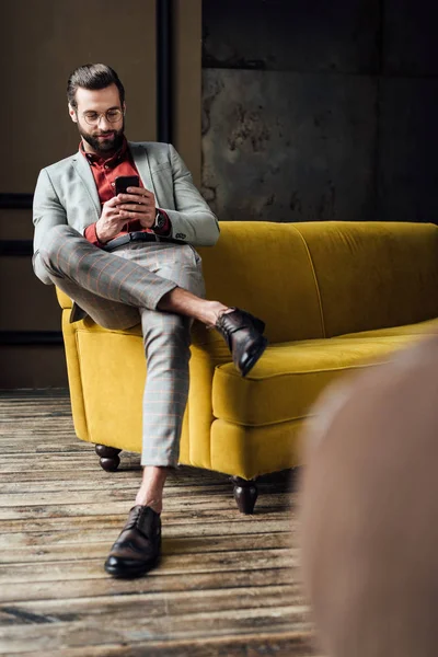 Hombre elegante guapo usando teléfono inteligente y sentado en el sofá amarillo — Stock Photo