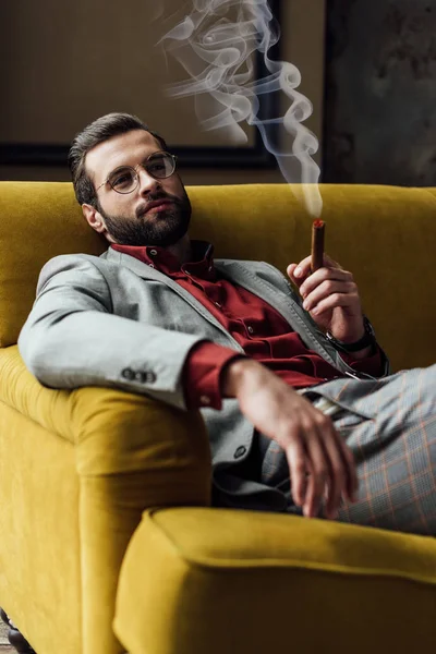 Красивий бородатий молодий чоловік курить сигару на дивані — стокове фото