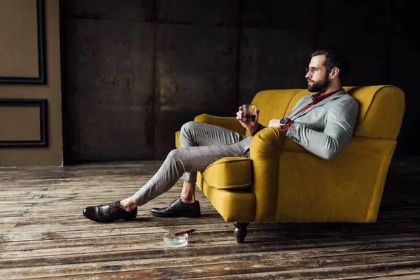 Стильный мужчина со стаканом виски сидит на диване с сигарой в пепельнице на полу — стоковое фото