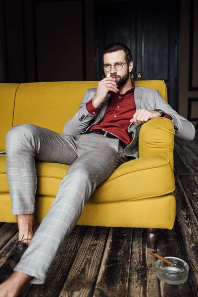 Bel homme avec un verre de whisky assis sur le canapé avec un cigare dans le cendrier sur le sol — Photo de stock