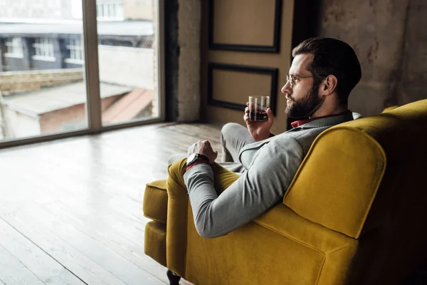 Uomo alla moda in possesso di un bicchiere di whisky e seduto sul divano — Foto stock