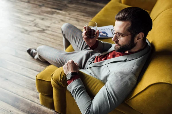 Homem elegante com uísque sentado no sofá com jornal — Stock Photo