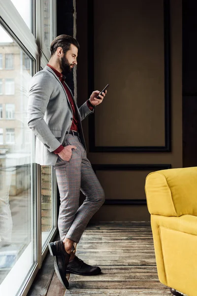 Homme à la mode en costume en utilisant smartphone à la fenêtre — Photo de stock