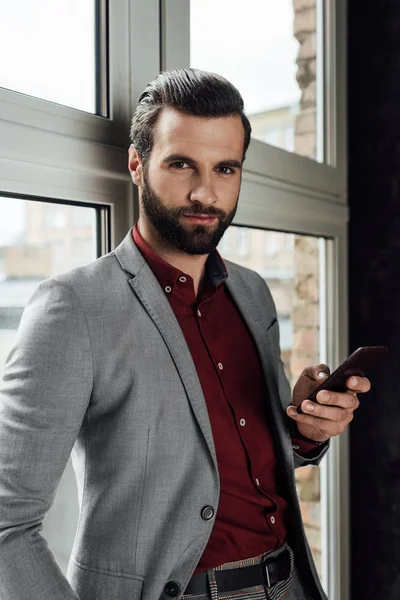 Hombre con estilo utilizando el teléfono inteligente en la ventana y mirando a la cámara - foto de stock