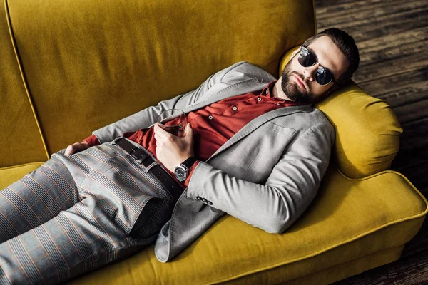 Homme ivre en lunettes de soleil avec verre de cognac dormant sur le canapé — Photo de stock