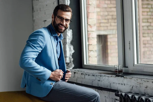 Hombre sonriente con estilo en gafas y traje azul de moda sentado en la ventana - foto de stock