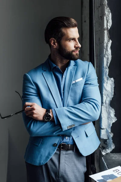 Модный вдумчивый мужчина в синем костюме держит очки и смотрит в окно — стоковое фото