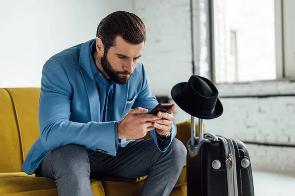 Hombre de negocios concentrado usando teléfono inteligente y sentado en el sofá con bolsa de viaje - foto de stock
