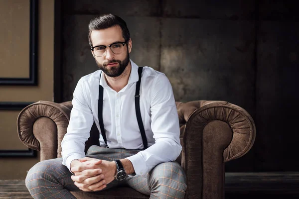 Bel homme élégant dans des lunettes et des bretelles assis dans un fauteuil — Photo de stock