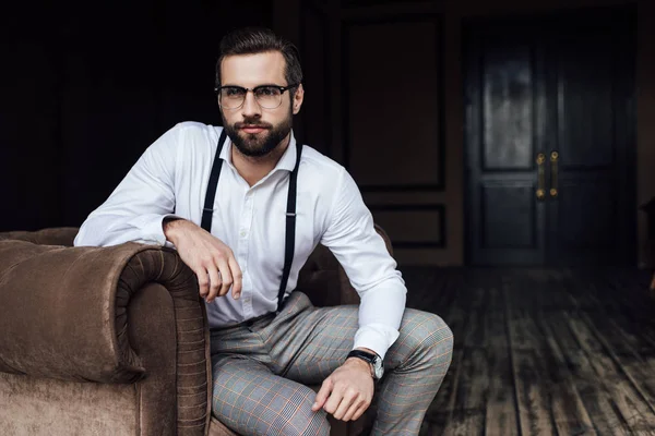 Homem barbudo na moda em óculos e suspensórios sentados em poltrona — Fotografia de Stock