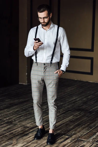 Stilvoller bärtiger Mann in Hosenträgern mit Smartphone im Loft-Inneren — Stockfoto
