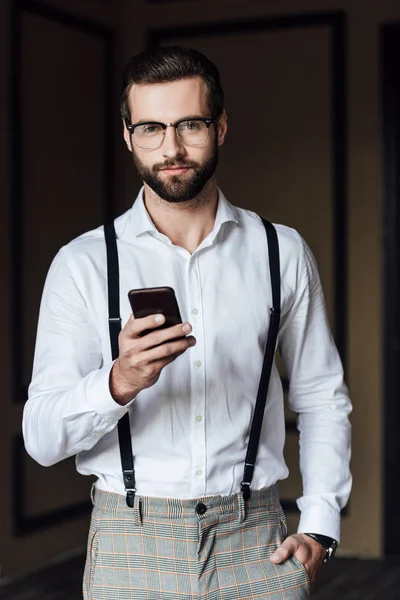 Bel homme barbu dans les bretelles en utilisant un smartphone et en regardant la caméra — Photo de stock