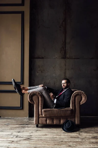 Homem de negócios elegante posando em poltrona, chapéu deitado no chão perto, loft interior — Fotografia de Stock