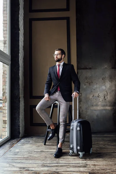 Elegant businessman in suit posing on stool with luggage — Stock Photo