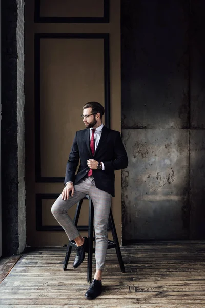 Schöner modischer Geschäftsmann im Anzug posiert auf einem Hocker im Loft-Interieur — Stock Photo