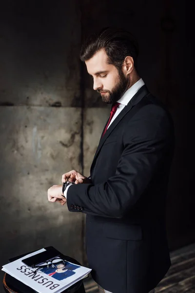 Bärtiger Geschäftsmann blickt auf Armbanduhr, während er am Hocker mit Wirtschaftszeitung und Smartphone steht — Stockfoto