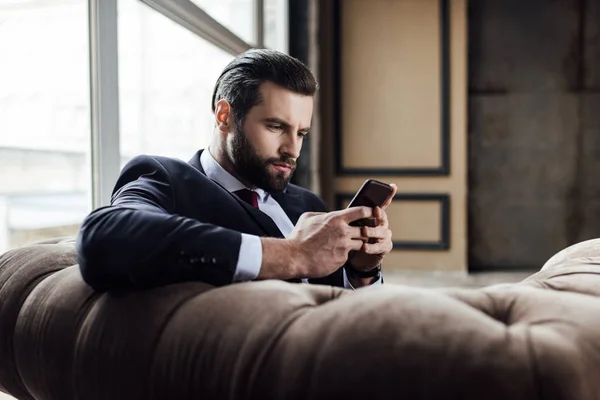 Hombre de negocios barbudo seguro usando teléfono inteligente y sentado en un sillón - foto de stock