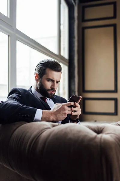 Модный бородатый бизнесмен, отправляющий сообщения на смартфон, сидя в кресле — стоковое фото