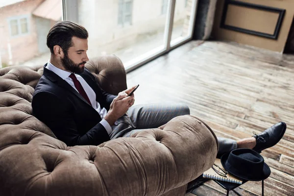 Confiado hombre de negocios elegante usando el teléfono inteligente y sentado en el sillón - foto de stock