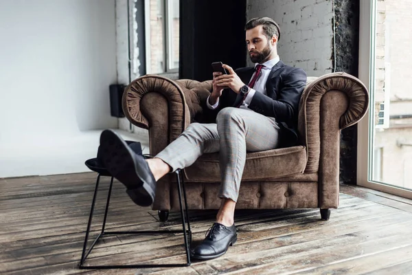 Hombre de negocios elegante usando el teléfono inteligente y sentado en el sillón en el loft - foto de stock