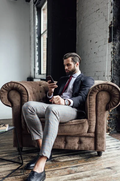 Homme d'affaires confiant textos sms sur smartphone tout en étant assis dans le fauteuil dans le grenier — Photo de stock
