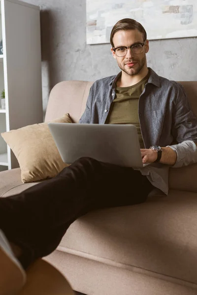Homme élégant travaillant avec un ordinateur portable sur le canapé et regardant la caméra — Photo de stock
