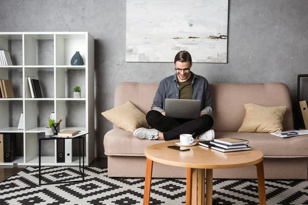 Jovem freelancer feliz trabalhando com laptop enquanto sentado no sofá — Fotografia de Stock