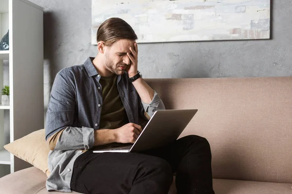 Junger überarbeiteter Mann mit Laptop hat Kopfschmerzen, während er auf der Couch sitzt — Stockfoto